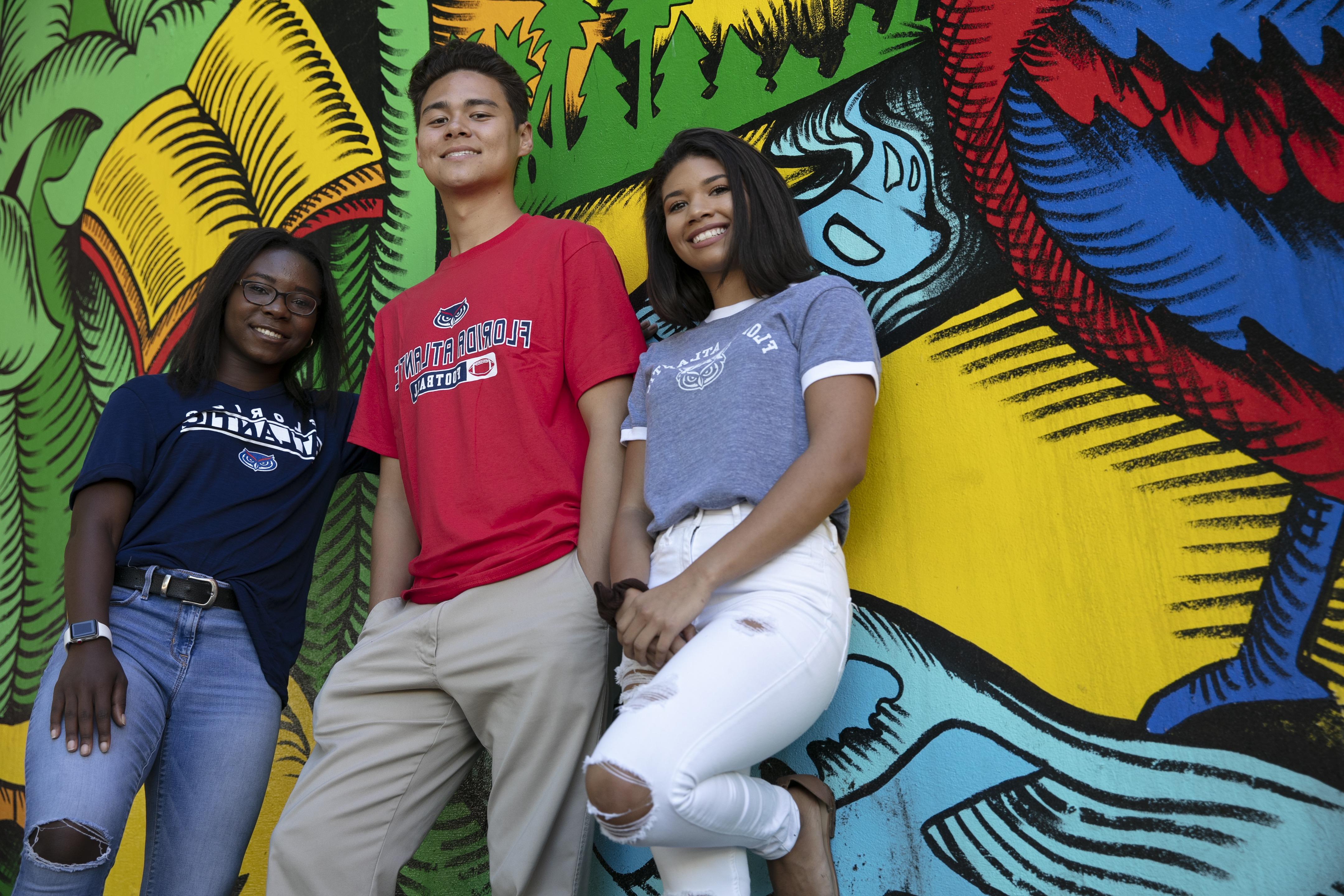 three students wearing FAU shirts stand in front of a mural while smiling at the camera